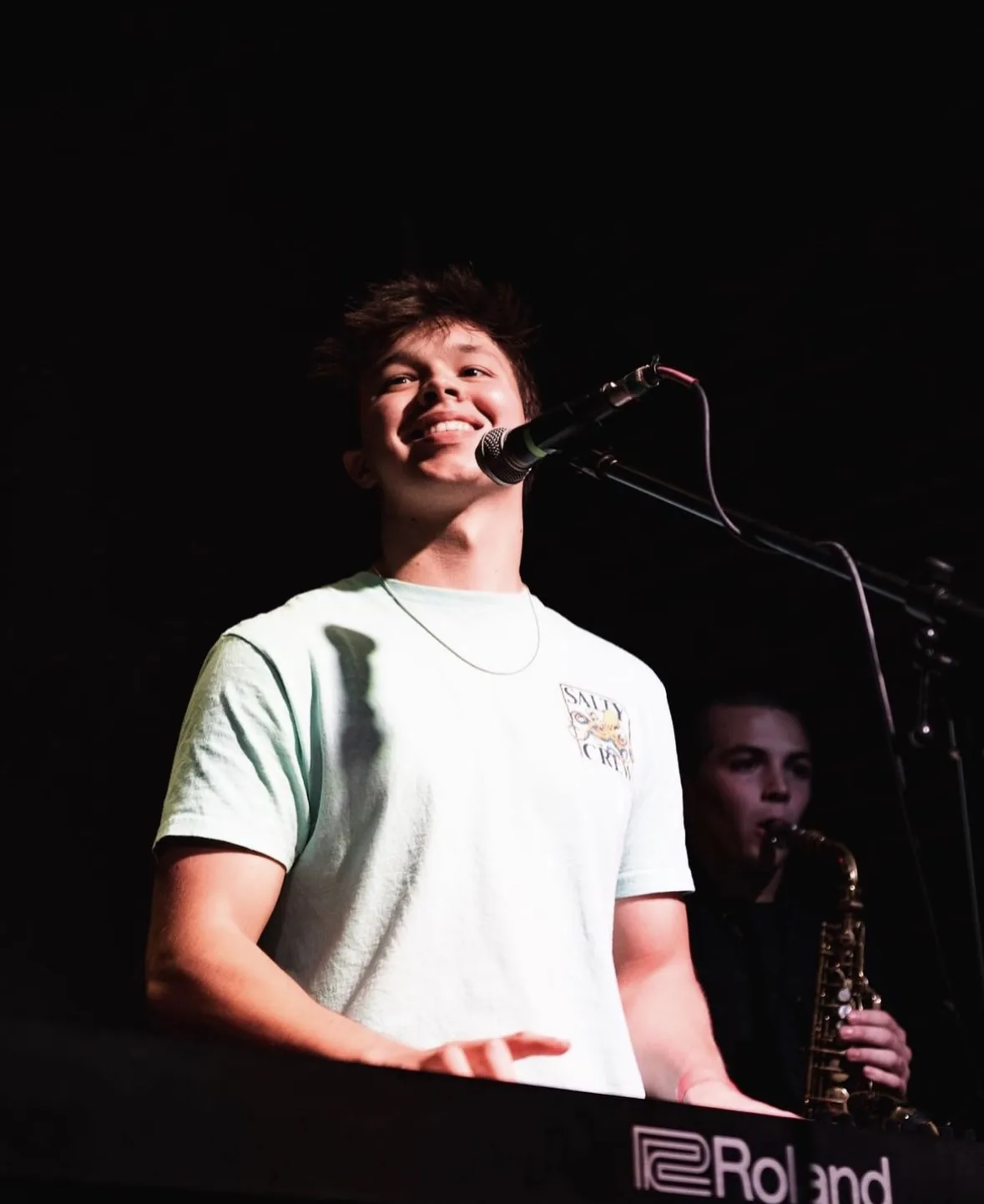 Max Cooper III smiling at the microphone mid-set, a saxophonist playing behind him